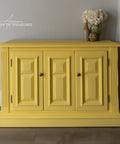 A bright yellow painted wooden sideboard cabinet with three paneled doors and small round knobs, shown in a styled room setting with a vase of flowers on top and a curtain to the right.
