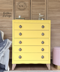 A bright yellow five-drawer chest of drawers with ornate silver knobs and light wood legs, styled in a bedroom setting against a wooden slatted wall.