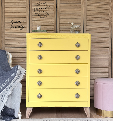 A bright yellow five-drawer chest of drawers with ornate silver knobs and light wood legs, styled in a bedroom setting against a wooden slatted wall.