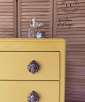 A close-up of a yellow painted dresser with ornate dark metal drawer pulls, set against a brown wooden shutter background. A seagull figurine and glass jar sit on top.