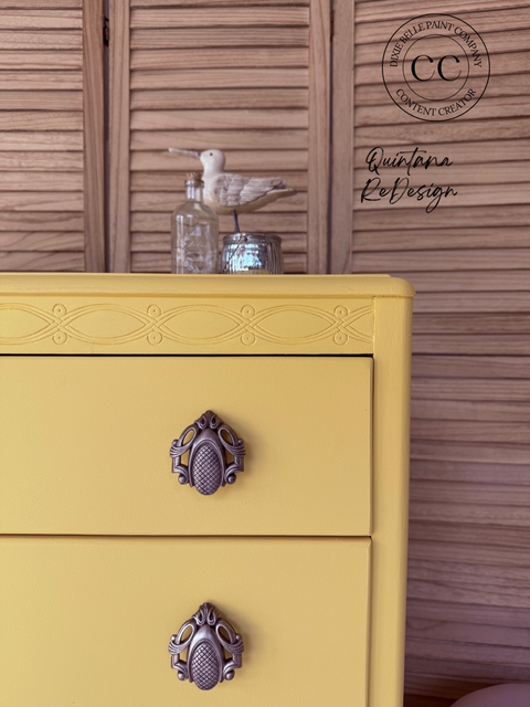 A close-up of a yellow painted dresser with ornate dark metal drawer pulls, set against a brown wooden shutter background. A seagull figurine and glass jar sit on top.