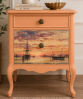 A peachy orange-colored nightstand with two drawers, featuring a detailed nautical sunset decoupage on the front of the lower drawer. The nightstand has cabriole legs, brass-style knobs, and is styled in a home interior setting with a plant and books. Label reads "Digital Rendering for Inspiration. Scale and sizing are approximate."