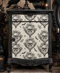 A gothic-style cabinet with a dark carved frame and a cream door covered in a black skull and ornate floral pattern, shown in a styled interior setting with candles and decorative objects.