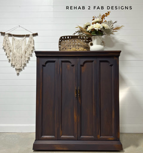 Dark wooden cabinet with decorative items against a white wall, featuring 'Rehab 2 Fab Designs' branding.