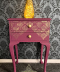 A purple two-drawer side table with gold damask-style painted details and gold knobs, styled with a glowing yellow decorative object on top, set against a gray damask wallpaper background.