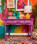A bright magenta-painted wooden console table styled with a yellow lamp, pink and blue pots, books, and decorative objects, set against a vividly painted multicolor wall.