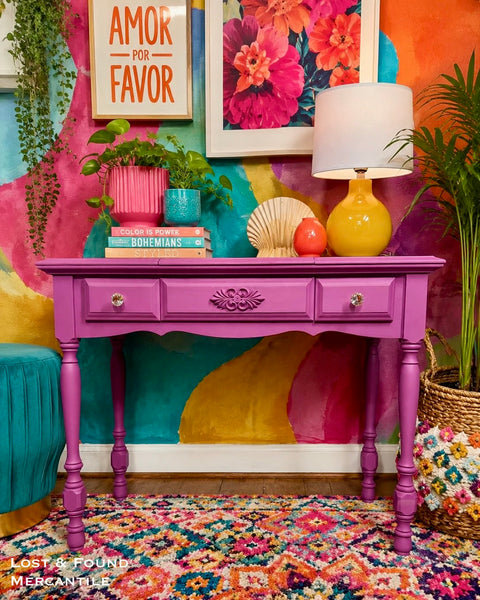 A bright magenta-painted wooden console table styled with a yellow lamp, pink and blue pots, books, and decorative objects, set against a vividly painted multicolor wall.