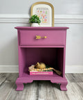 A purple nightstand with a single drawer and an open lower shelf, styled with a gold arrow drawer pull, a small potted plant, and decorative items on a wood floor against a white wall.