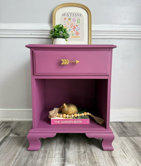 A purple nightstand with a single drawer and an open lower shelf, styled with a gold arrow drawer pull, a small potted plant, and decorative items on a wood floor against a white wall.