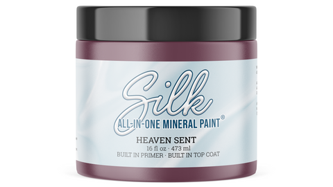 A jar of Silk All-In-One Mineral Paint in the color Heaven Sent, featuring a dark plum-purple container, a black lid, and a light blue and white label.