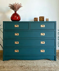 Teal dresser with gold handles against a white wall, featuring a red vase and decorative items.