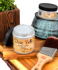 Dixie Belle glaze jars and a brush on a wooden surface with a plant in the background