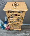 A painted wooden jewelry chest with a warm beige and orange checkerboard pattern, featuring floral designs on the drawer fronts and a dark damask motif on the top surface. The chest is styled on a gray wood floor against a gray wall, with decorative dried flowers on the left.