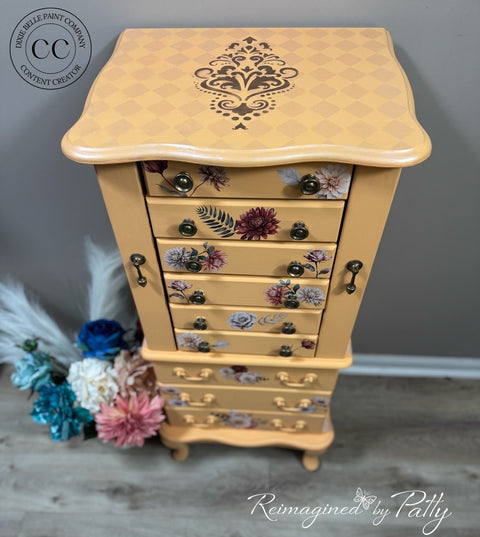 A painted wooden jewelry chest with a warm beige and orange checkerboard pattern, featuring floral designs on the drawer fronts and a dark damask motif on the top surface. The chest is styled on a gray wood floor against a gray wall, with decorative dried flowers on the left.