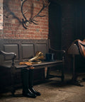 Rustic dark wood bench styled with riding boots, leather gloves, and a brass horn in a moody equestrian-inspired room with exposed brick walls and a saddle display.