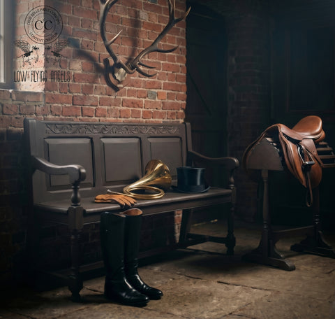 Rustic dark wood bench styled with riding boots, leather gloves, and a brass horn in a moody equestrian-inspired room with exposed brick walls and a saddle display.