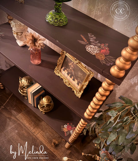 Decorative shelf with various items including a gold frame and a green vase, with 'by Melinda' branding.