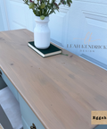 A light natural wood vanity top or desk surface shown in a real interior setting, with a white vase and book on top. The scene includes a white paneled door in the background and a visible Leah Kendrick Design watermark; a small label in the corner reads Eggshell.
