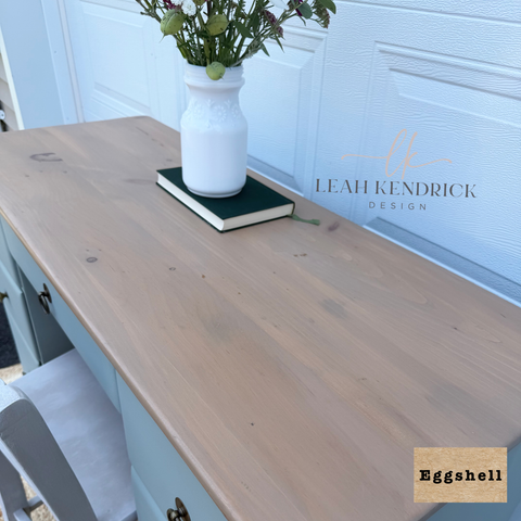A light natural wood vanity top or desk surface shown in a real interior setting, with a white vase and book on top. The scene includes a white paneled door in the background and a visible Leah Kendrick Design watermark; a small label in the corner reads Eggshell.