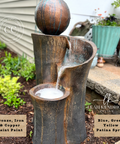 A decorative outdoor water fountain with a spherical top, shown with water flowing into a basin. The product has a distressed bronze and iron finish with blue, green, and yellow patina spray accents.
