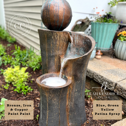 A decorative outdoor water fountain with a spherical top, shown with water flowing into a basin. The product has a distressed bronze and iron finish with blue, green, and yellow patina spray accents.