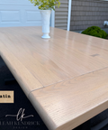 A light oak extendable dining table with a satin finish, shown outdoors with a white vase on top. A wood swatch labeled 'Satin' and the brand 'Leah Kendrick Design' are visible in the lower left corner.