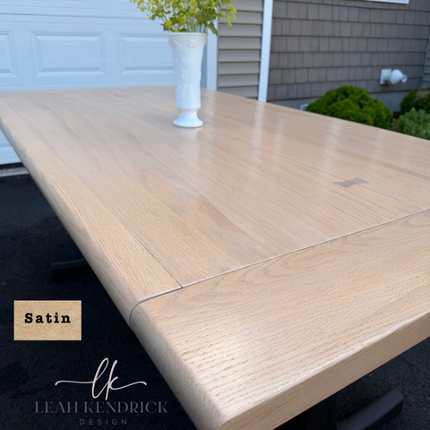 A light oak extendable dining table with a satin finish, shown outdoors with a white vase on top. A wood swatch labeled 'Satin' and the brand 'Leah Kendrick Design' are visible in the lower left corner.