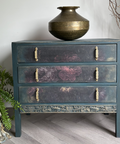 Vintage dresser painted in a deep moody blue finish with floral detailing on the drawers, gold hardware, and carved accents, styled with greenery and antique-inspired decor.