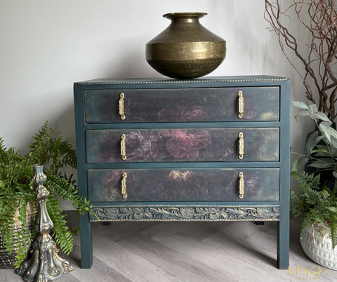 Vintage dresser painted in a deep moody blue finish with floral detailing on the drawers, gold hardware, and carved accents, styled with greenery and antique-inspired decor.