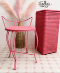 A pink metal chair with a round seat and curved legs stands beside a tall pink woven storage bin against a white tile wall. Pink pampas grass in a woven vase sits behind the table, and a 'Tracey's Fancy' logo is visible on the wall.