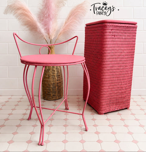 A pink metal chair with a round seat and curved legs stands beside a tall pink woven storage bin against a white tile wall. Pink pampas grass in a woven vase sits behind the table, and a 'Tracey's Fancy' logo is visible on the wall.