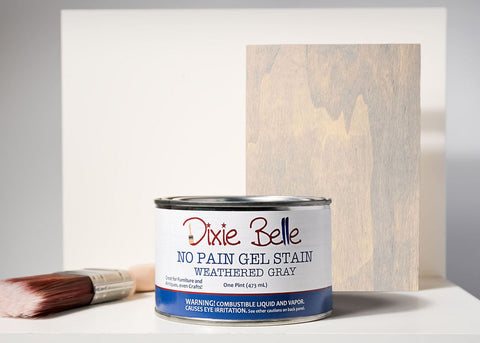 Can of Weathered Gray Dixie Belle gel stain with a brush and stained wood panel on a gray background