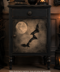 A dark vintage-style cabinet or nightstand with a distressed black finish, featuring a door panel decorated with a full moon and three silhouetted bats. The cabinet stands in a dimly lit room with candles and wood paneling in the background.