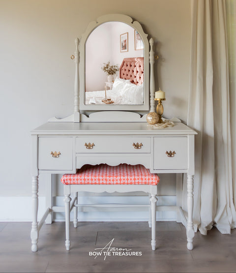 Vintage-style vanity with mirror and chair in a room with curtains and a window.