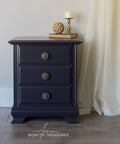 Black nightstand with three drawers against a neutral wall with a white curtain.