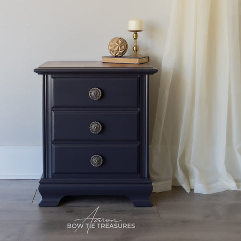 Black nightstand with three drawers against a neutral wall with a white curtain.