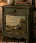 A vintage-style painted cabinet shown in a styled interior scene. The cabinet is dark green with ornate turned legs, a single drawer with a round knob, and a front panel featuring a painted pastoral landscape with cows and trees.