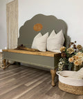 a long bench that is painted in all in one paint in a forest green color, two white pillows and a bundle of straw are staged on the bench. to the left of the bench is a wall handing made of rope and lots of plants in containers to the right of the bench.  Also a basket with a rug to the right.