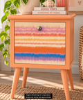 A peachy orange-colored wooden nightstand with a colorful striped decoupage on the front, shown in a styled room setting with a plant, books, and a woven chair. Label reads "Digital Rendering for Inspiration. Scale and sizing are approximate."
