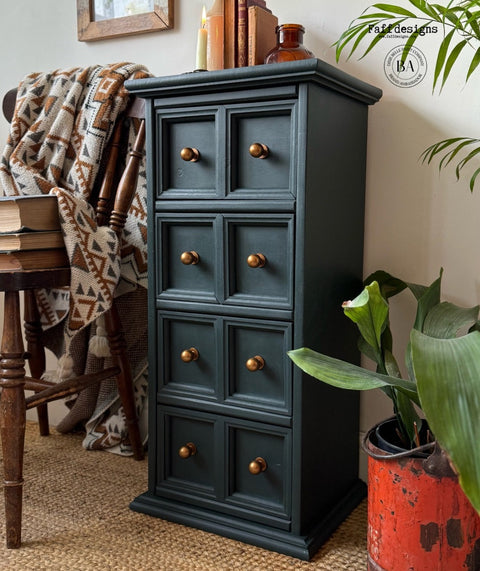 Dark green wooden cabinet with brass knobs in a home setting with a plant and books.