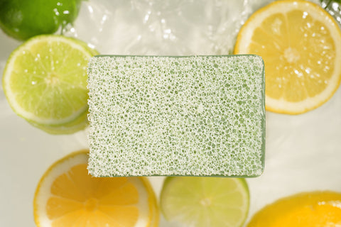 Lemon line Scrubby soap with suds surrounded by sliced lemons and limes on a white surface