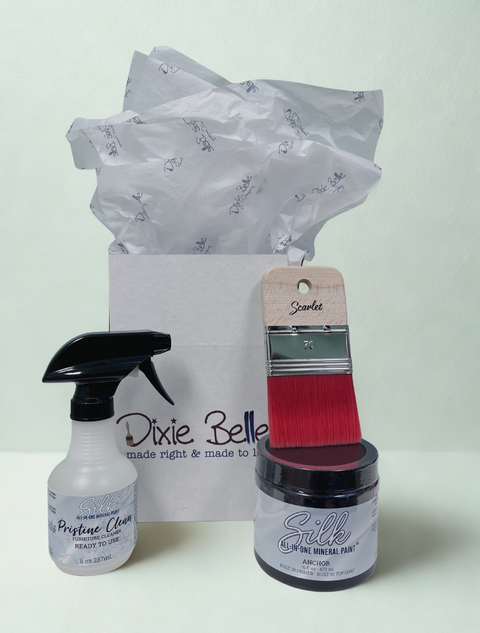 A Dixie Belle gift set featuring a bottle of Pristine Clean Ready to Use, a jar of Silk Paint in Anchor, and a paintbrush, arranged in front of a branded white gift bag with tissue paper.