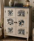 A vintage-style painted cabinet with six doors, featuring blue-toned illustrated scenes on an off-white distressed finish, styled in a cozy room setting with a blue top, books, a lantern, and potted plants.