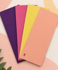 Four rectangular paper swatches in purple, pink, yellow, and orange, arranged in a fan shape on a light background with soft pink and yellow abstract shapes and a small green leaf at the lower left.
