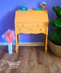 An orange vintage writing desk styled in a room with a purple wall and wood floor, decorated with small figurines and framed art.