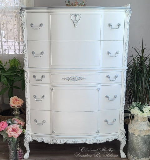 Decorative white dresser with intricate details in a room with plants and flowers.