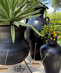 Three black decorative ceramic pots in different shapes, shown outdoors with plants. The pots are labeled 'Eggshell', 'Satin', and the image includes the brand name 'Tracey's Fancy' at the bottom.
