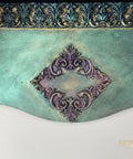 An ornate decorative wall panel or wallpaper sample featuring a distressed teal-green background, a central purple damask medallion, and a metallic bronze-accented top border. The brand name 'MCKAY DESIGNS' is visible in the bottom right corner.