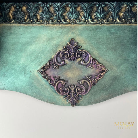 An ornate decorative wall panel or wallpaper sample featuring a distressed teal-green background, a central purple damask medallion, and a metallic bronze-accented top border. The brand name 'MCKAY DESIGNS' is visible in the bottom right corner.