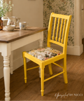 A bright yellow wooden dining chair with a floral-patterned cushion sits next to a white table with a wooden top in a cozy, vintage-style room with floral wallpaper and wood floors.